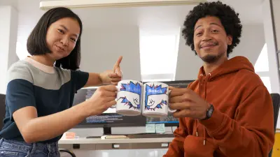 Junge Frau und Mann lächeln mit Daumen nach oben und stoßen mit Bechern an.