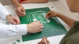 Sarah, Simon und Mustafa haben jeweils ein Puzzleteil in der Hand. Ein fast fertiges Puzzle liegt auf einem Tisch. Alle legen ihr Teil in die passenden Lücken und das Puzzle wird vervollständigt.